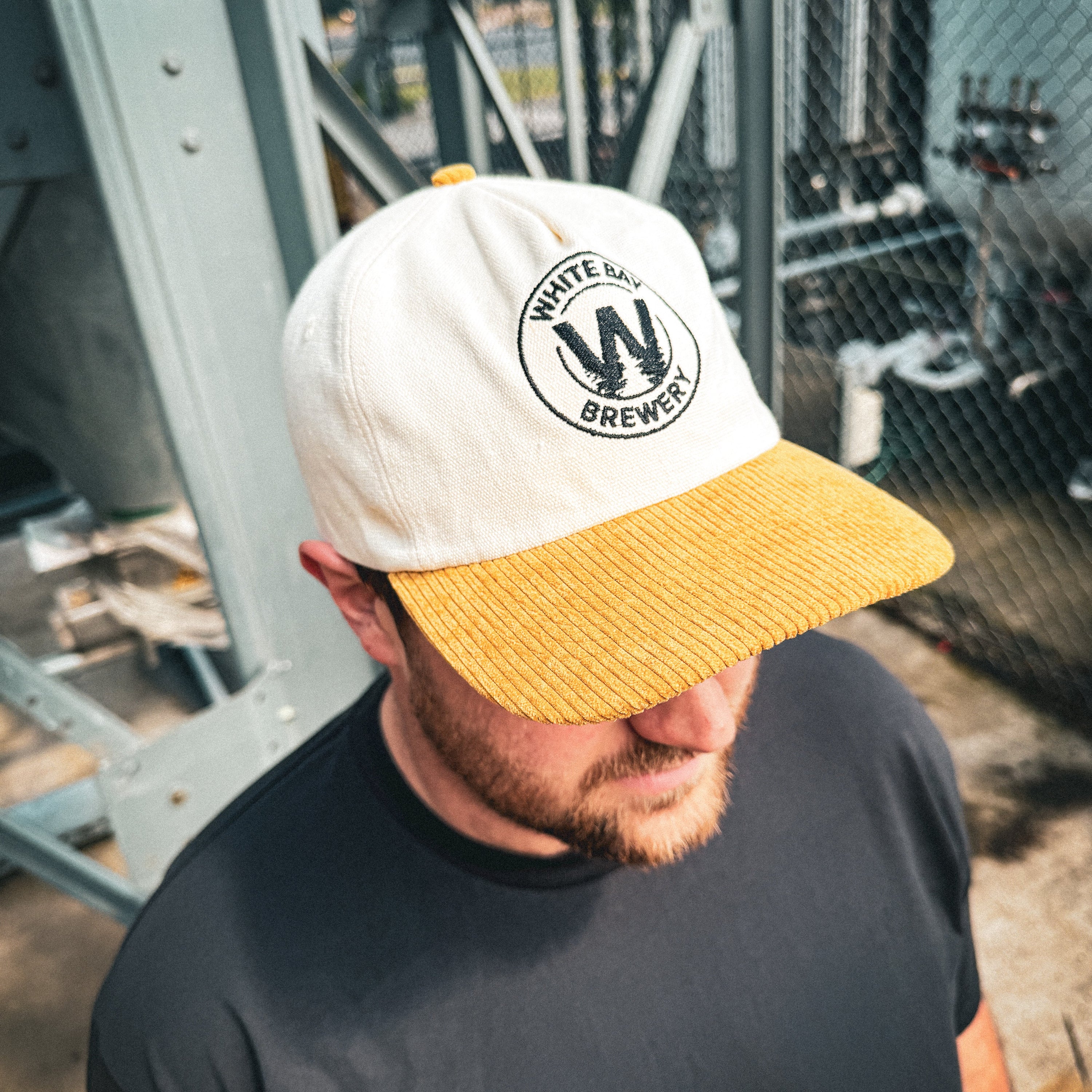Man wearing a white and yellow cap with a logo, standing in an industrial setting.
